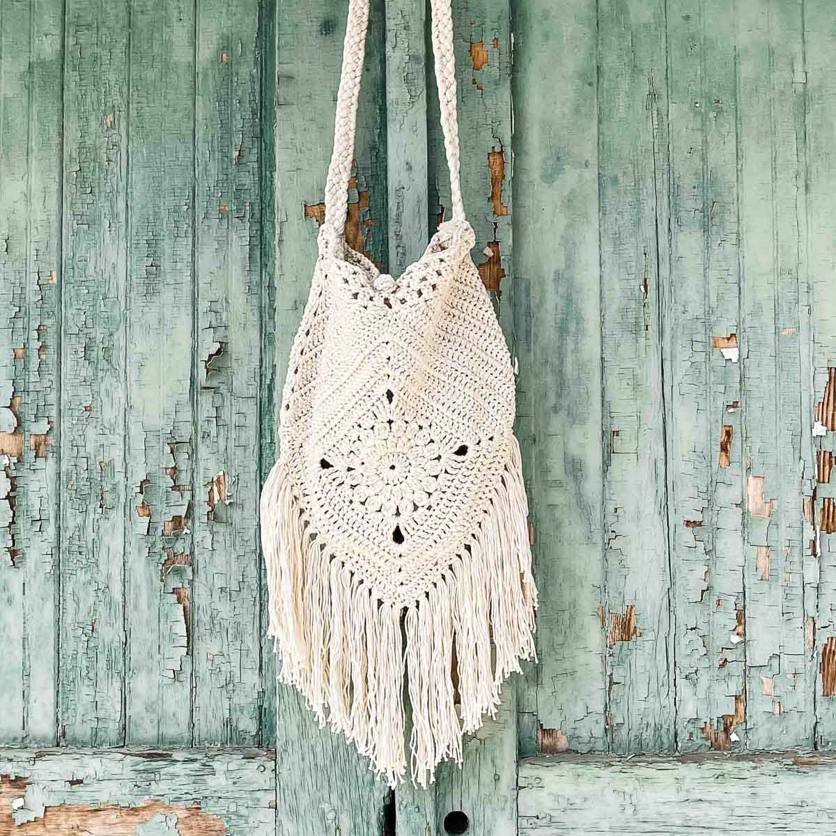 White boho crochet bag with long fringe hanging on weathered turquoise wooden wall — handmade summer purse with tassels