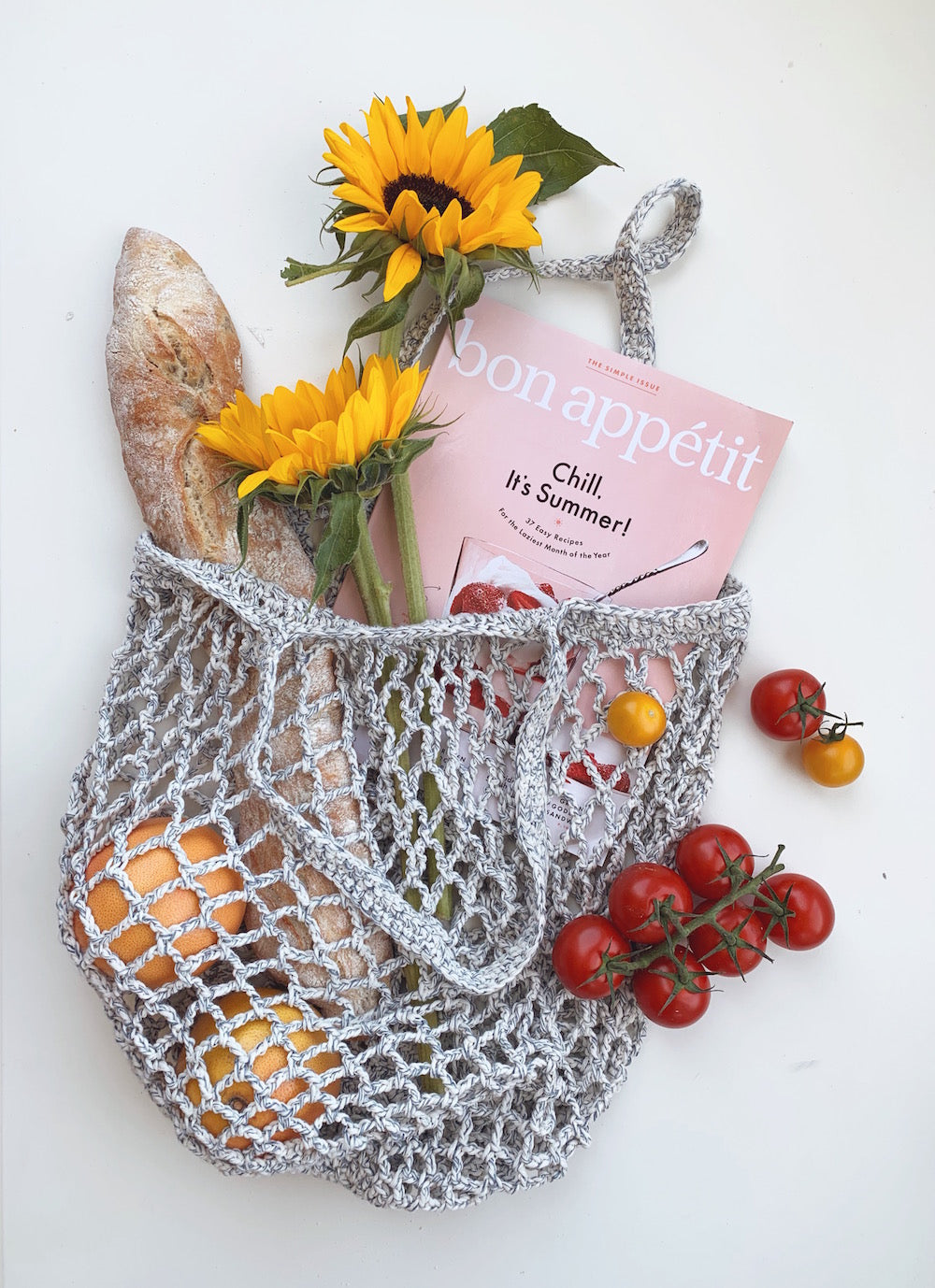 Grey crocheted market bag flat lay with a baguette, sunflowers, oranges, cherry tomatoes and a magazine